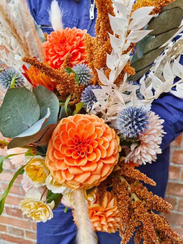 Bridal bouquet in rustic, autumnal colours. With eucalyptus, orange dahlias, globe thistles, ruskus, roses, and grasses. A mix of dried and fresh flowers.