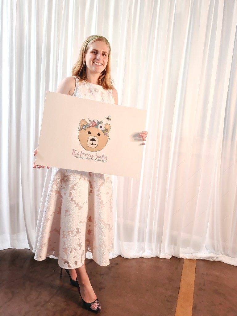 Two brides holding a wedding bouquet and smiling at the camera, a logo for The Honey Seeker Floral Design featuring the bear with a flower crown and a bee, and Esme Ford holding her sign at the wedding fair.