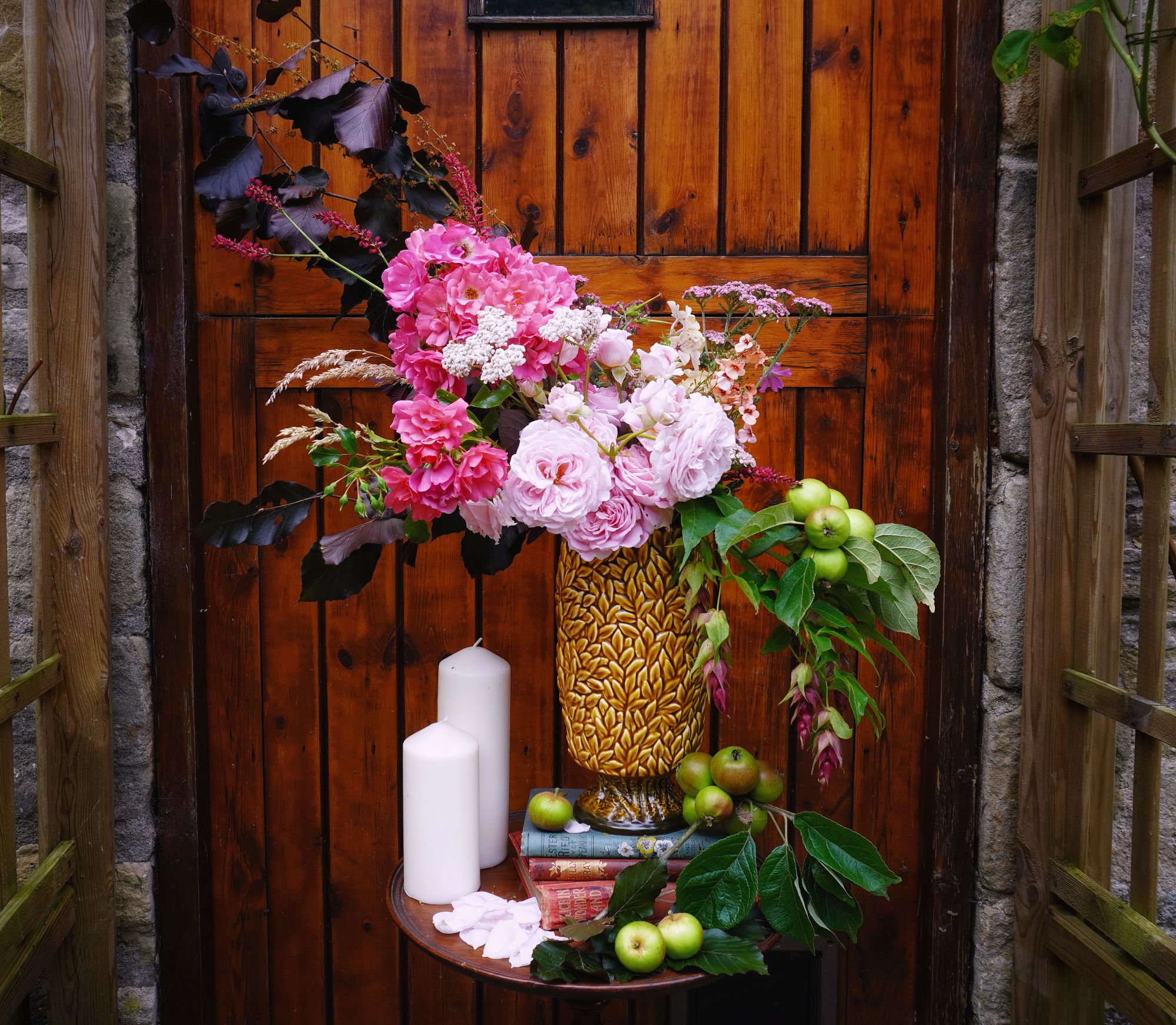 Lavish floral arrangement, or centrepiece, in a decadent botanical vase in mustard colour and leaf motives. Filled with bold pink roses, wild meadow flowers, purple leaves, and apples. On a stand of books, with candles.
