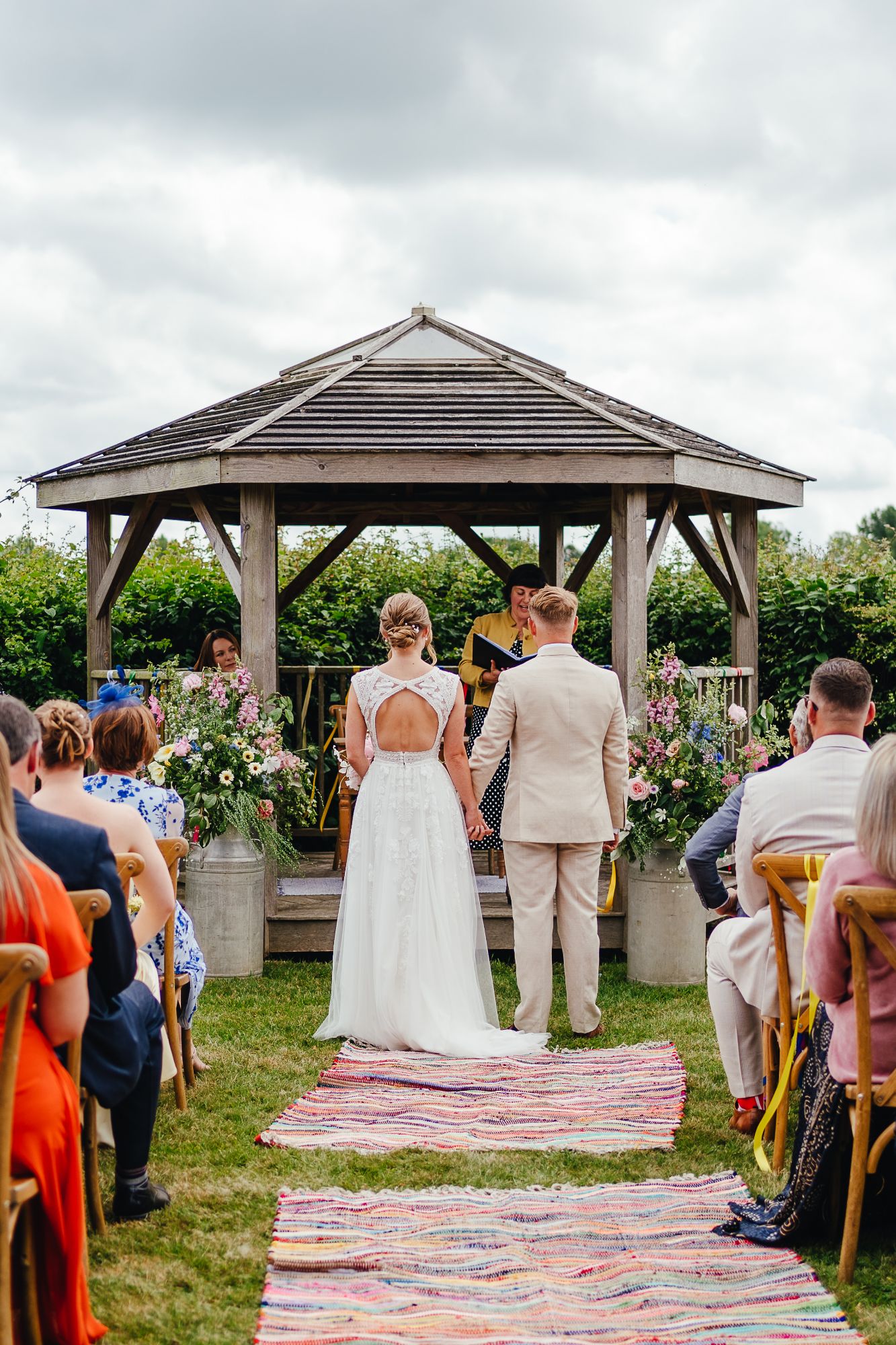 Couple at the wedding altar, surrounded by flowers at Pennard Hill Farm in Somerset. Boho, colourful wedding, with milk church flowers by The Honey Seeker Floral Design. Image by Freya Steele.