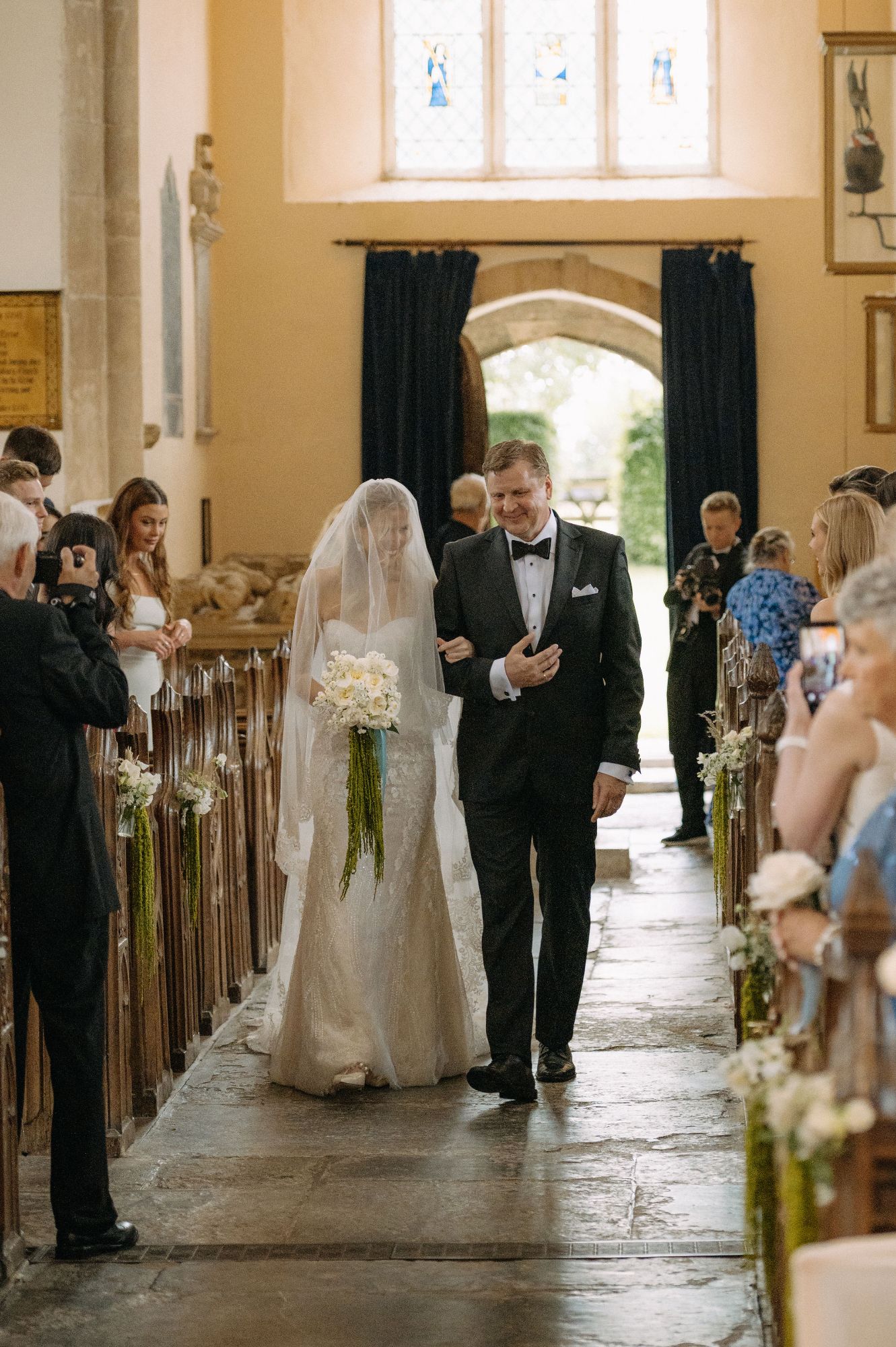 Bride walking down the church aisle of St. Michael's at North Cadbury Court, with her father. Holding flowers arranged by The Honey Seeker Floral Design in Somerset, with aisle flowers and sweetpeas lining the pews. Photo: Sophia Veres