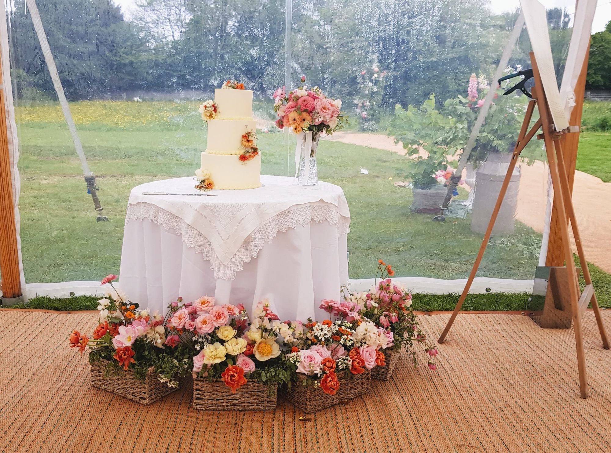 Wedding cake table flowers - meadow boxes in corals, yellows, pinks, whites, surrounding a simple 3 tier wedding cake in a marquee